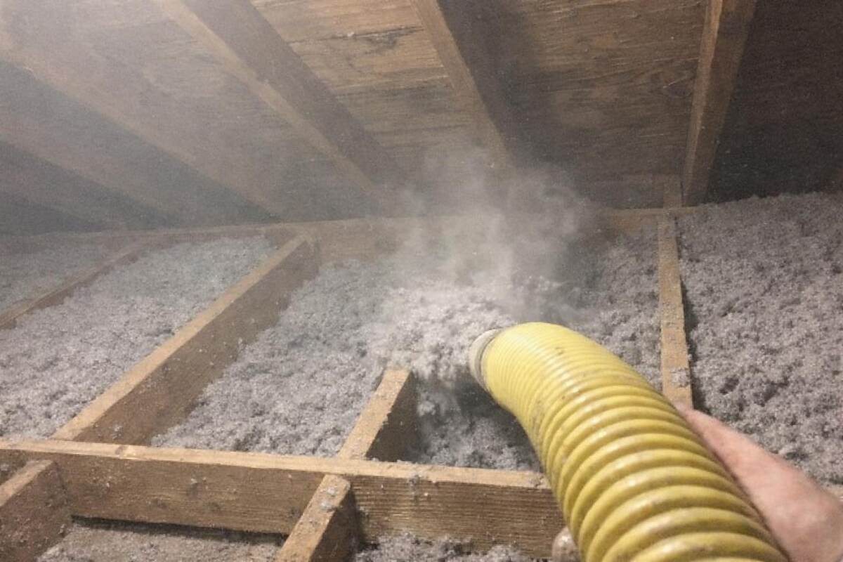 A blown-in insulation hose filling a joist bay with loose gray insulation in a low attic